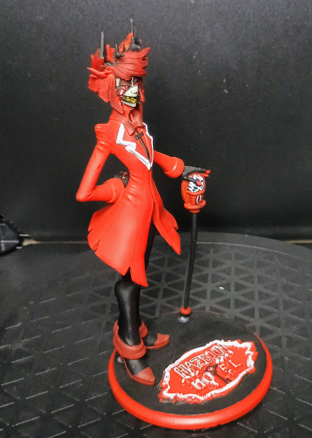 A red devil-themed figurine standing on a circular base with a cane, photographed inside a black display case or box.